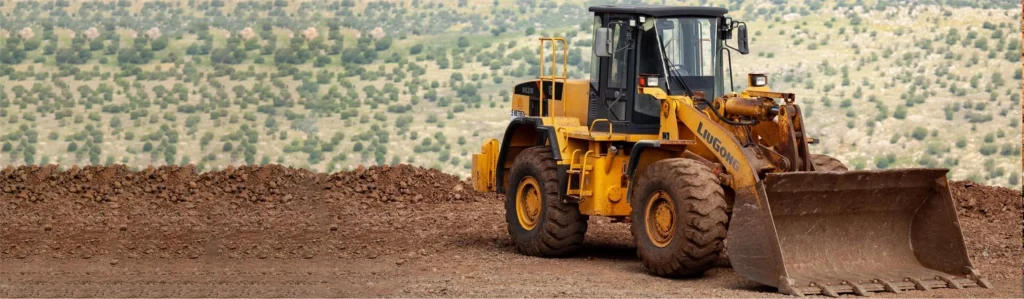 A large yellow LiuGong wheel loader standing on a dirt mound at a construction site with a hilly, green landscape in the background.