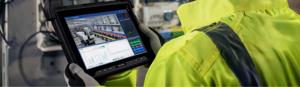 An engineer in a bright yellow jacket holding a rugged tablet that shows a live video feed of a production line and technical performance graphs.