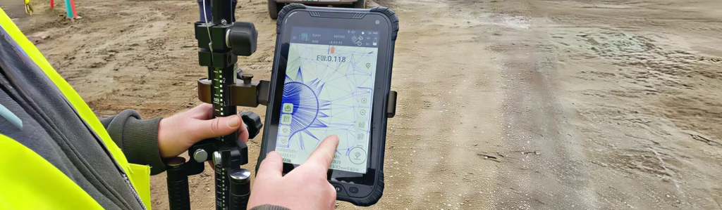 Close-up of a surveyor’s hands using a rugged tablet mounted on a surveying pole to view a digital mapping interface on a dirt construction site.