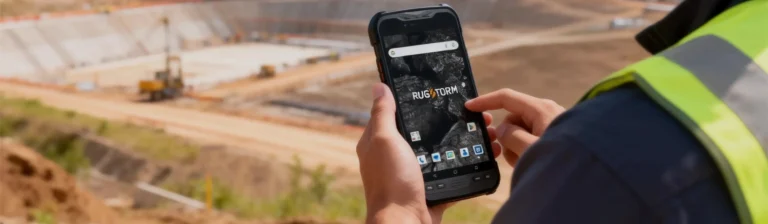 A person holding a RUGSTORM rugged smartphone with a reinforced casing overlooking a large-scale excavation and construction site.