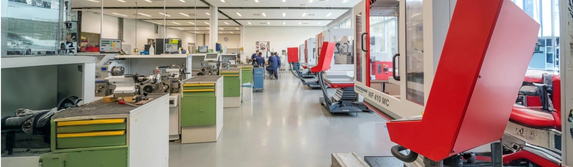 A panoramic view of a clean, modern precision machining workshop. On the right, large red and white CNC milling machines are aligned, including a DECKEL MAHO WF 410 MC. On the left, there are workbenches, manual lathes, and green and yellow tool cabinets, with two technicians working in the distance.