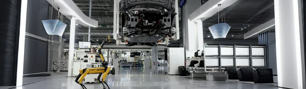 A Boston Dynamics Spot robot dog standing in a modern, brightly lit automotive manufacturing plant with a car chassis suspended on an assembly line.