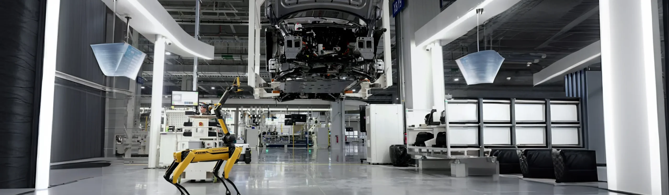A Boston Dynamics Spot robot dog standing in a modern, brightly lit automotive manufacturing plant with a car chassis suspended on an assembly line.