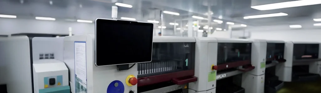 A black touch screen monitor with a silver frame mounted on industrial machinery in a clean factory, shown with a dark standby screen.
